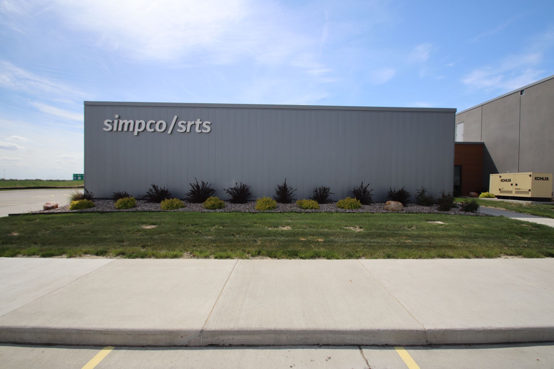 Gray industrial building with “simco/srts” sign, small shrubs, grass, and a sidewalk in front