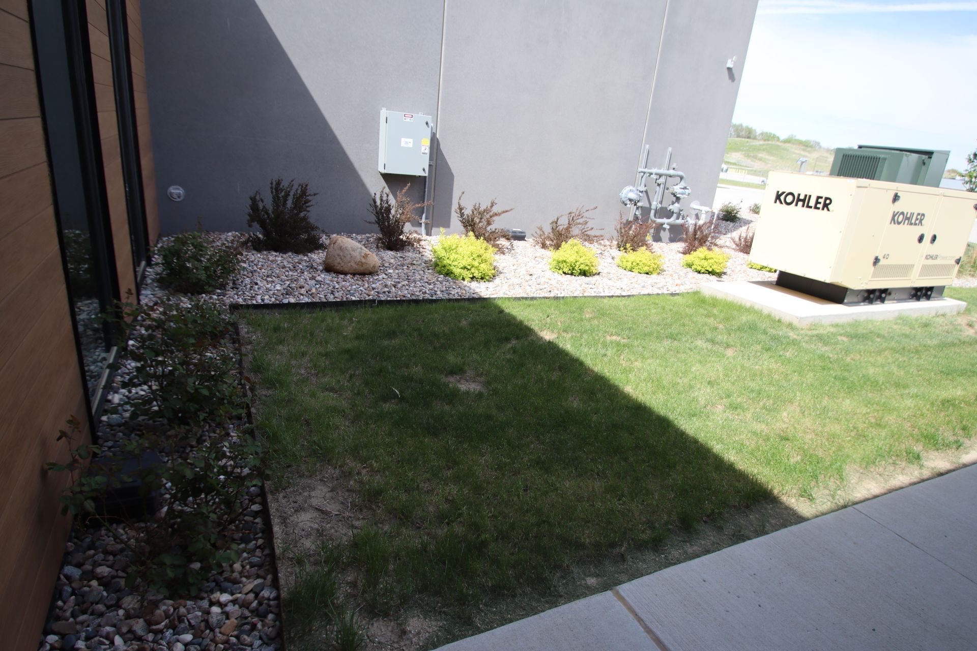 Small grassy courtyard beside a gray building, with rocks, yellow flowers, and an HVAC unit.