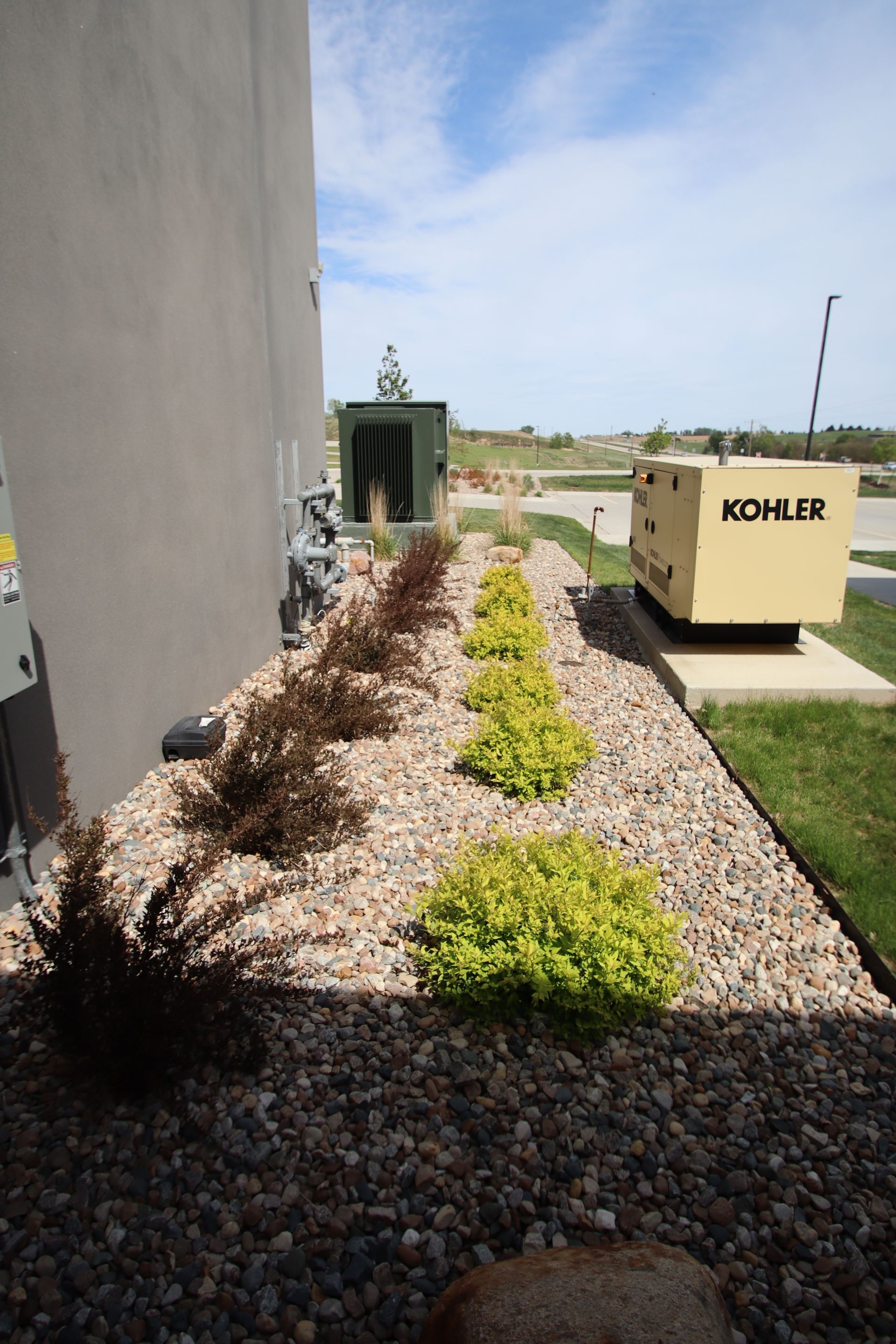 Landscaped gravel strip beside a building, with small shrubs and a beige utility box labeled “KOHLER.”