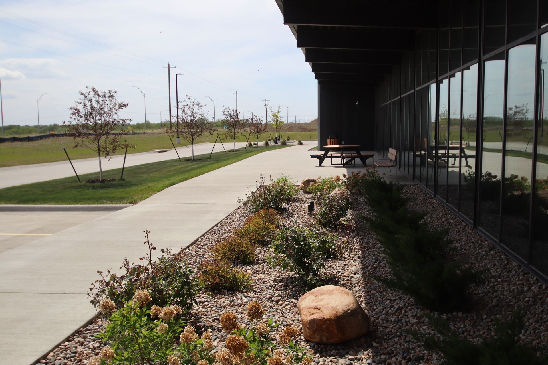 Sidewalk beside a building with glass windows, picnic tables, and a landscaped planting bed under a clear sky.