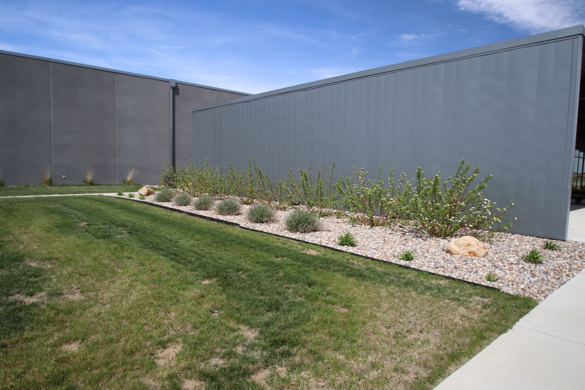Landscaped corner with gray walls, green grass, and a gravel bed of small plants under a blue sky.