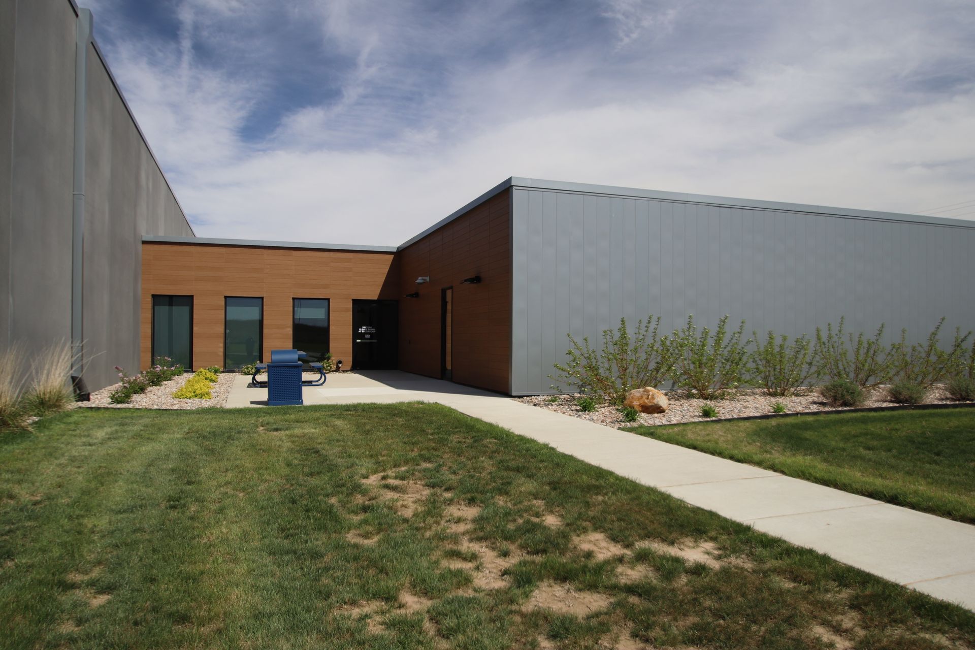 Modern building exterior with a walkway, grass, and a blue trash bin under a cloudy sky