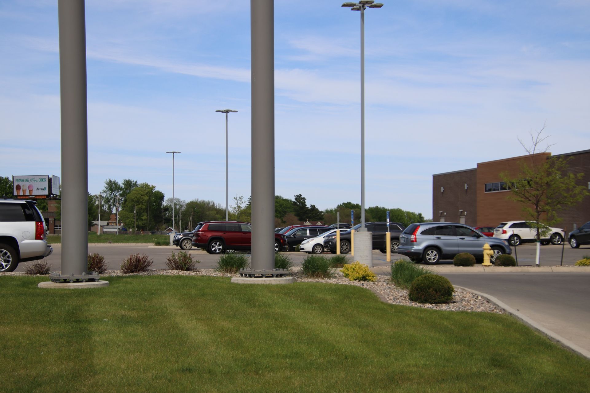 Parking lot with cars and tall light poles beside a brick building and grassy median
