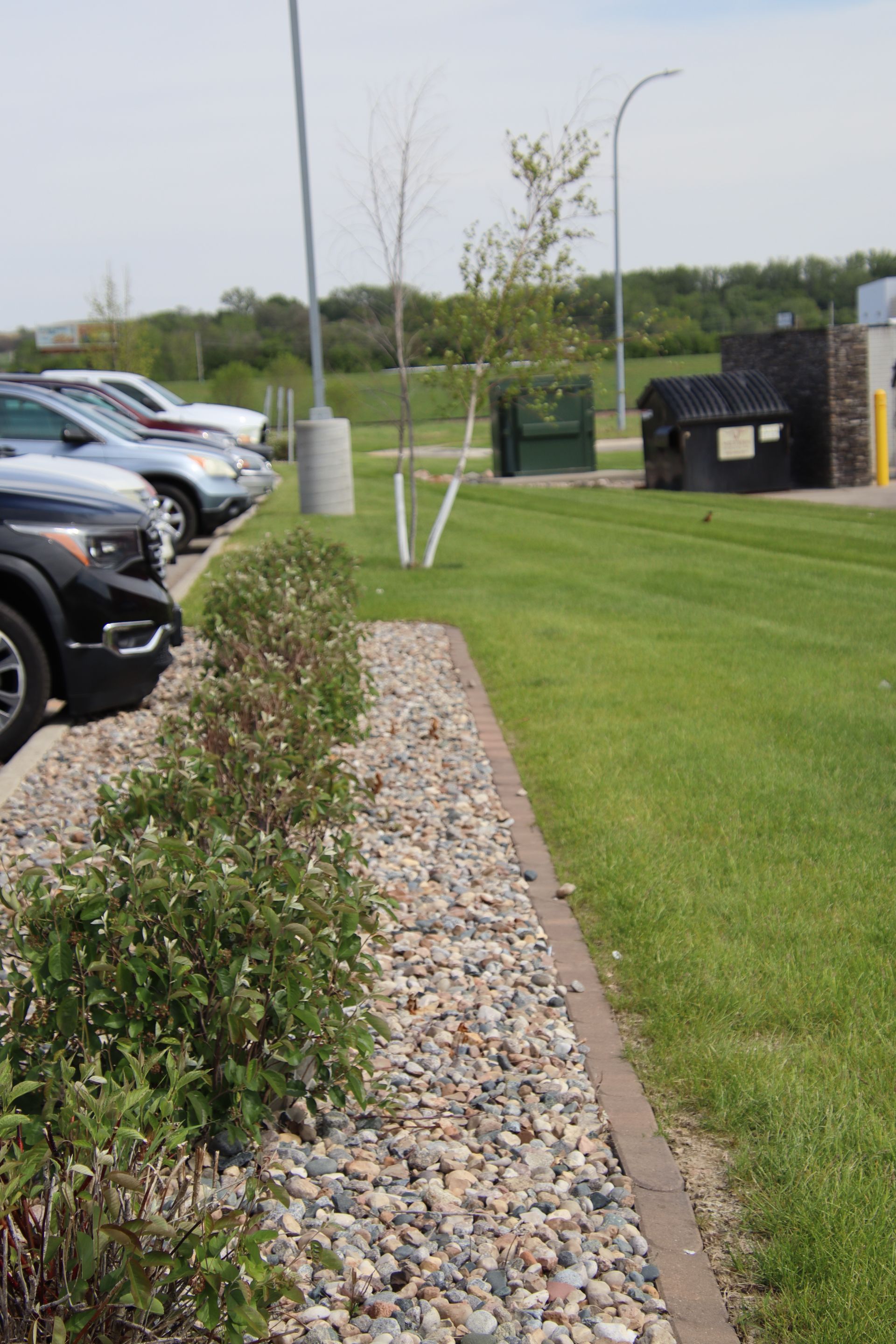 Parking lot with cars, a gravel bed, trimmed grass, and small trees beside a sidewalk near a brick structure