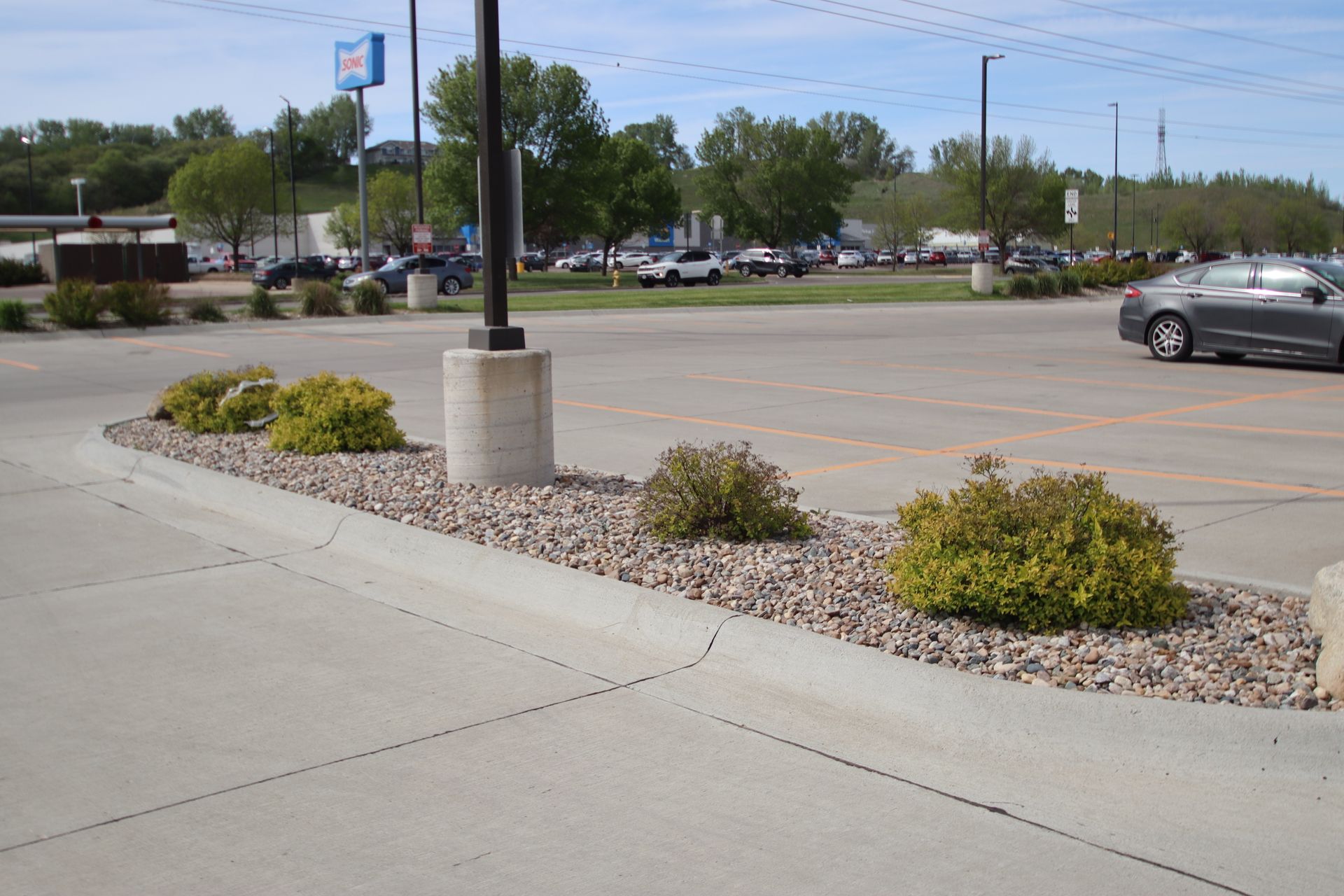 Landscaped parking lot with concrete sidewalk, rock bed, shrubs, and parked cars under a clear sky