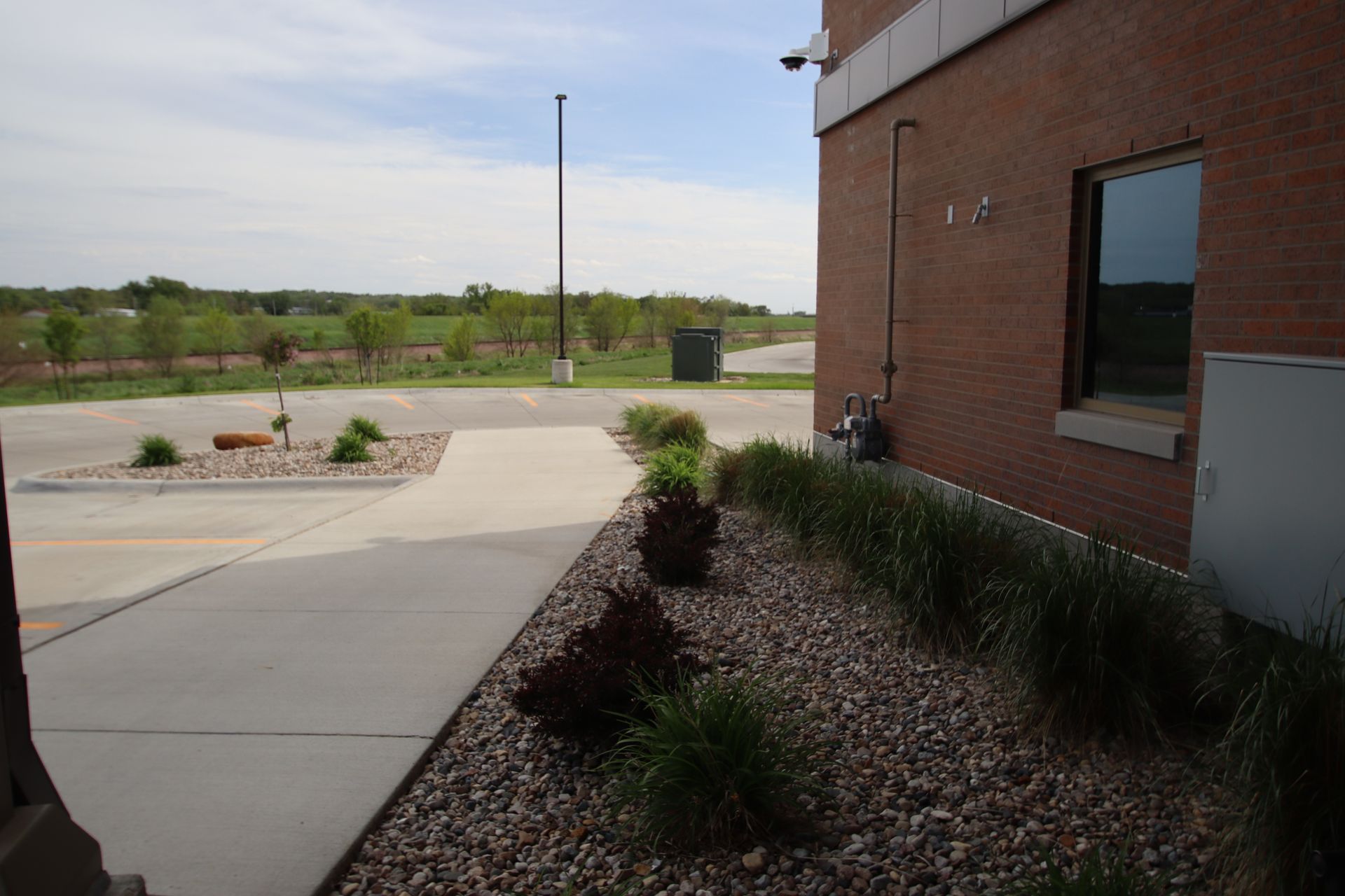 Brick building beside a sidewalk and landscaped gravel beds, with open field and cloudy sky beyond