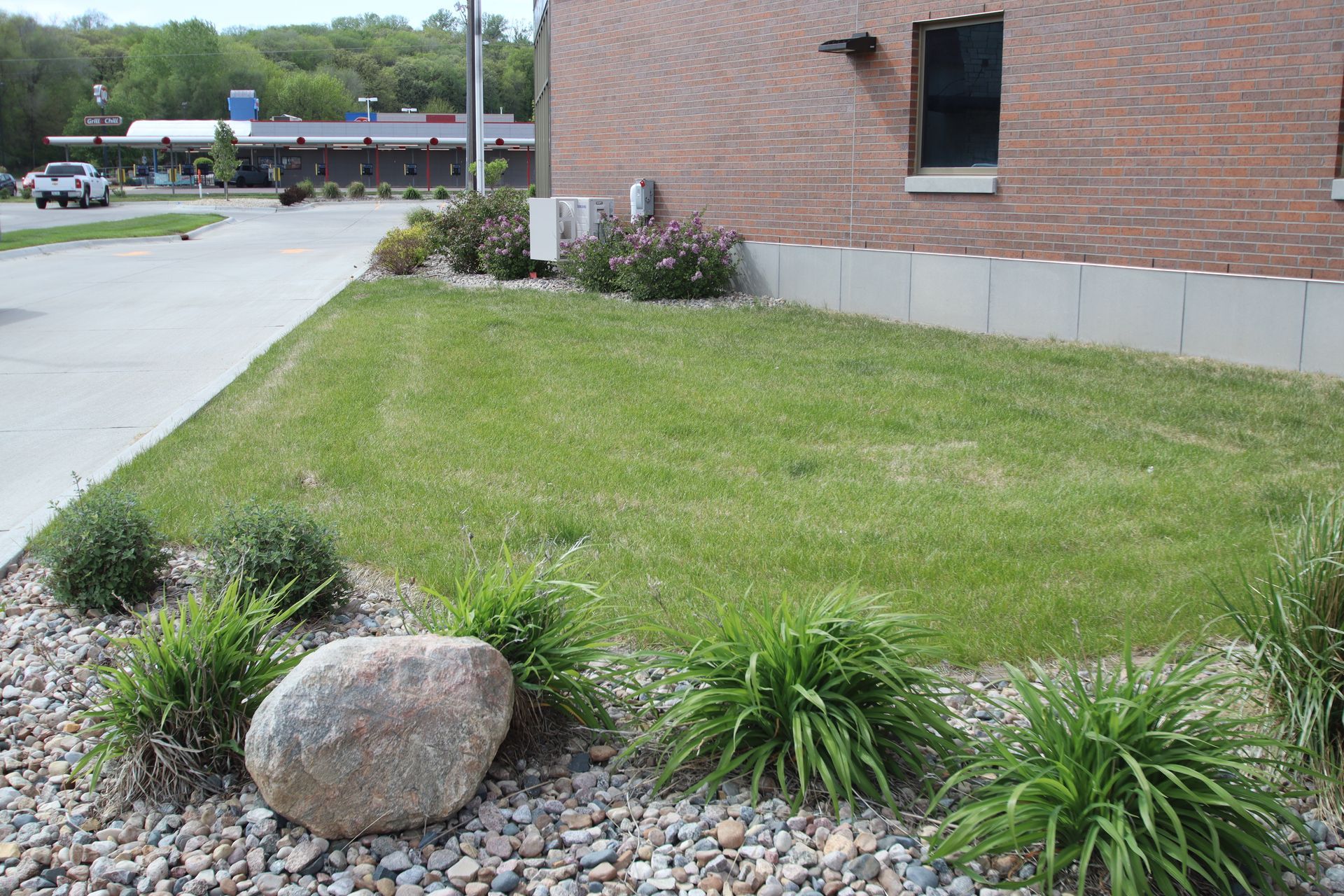 Landscape bed with grass, rocks, and shrubs beside a brick building and parking lot