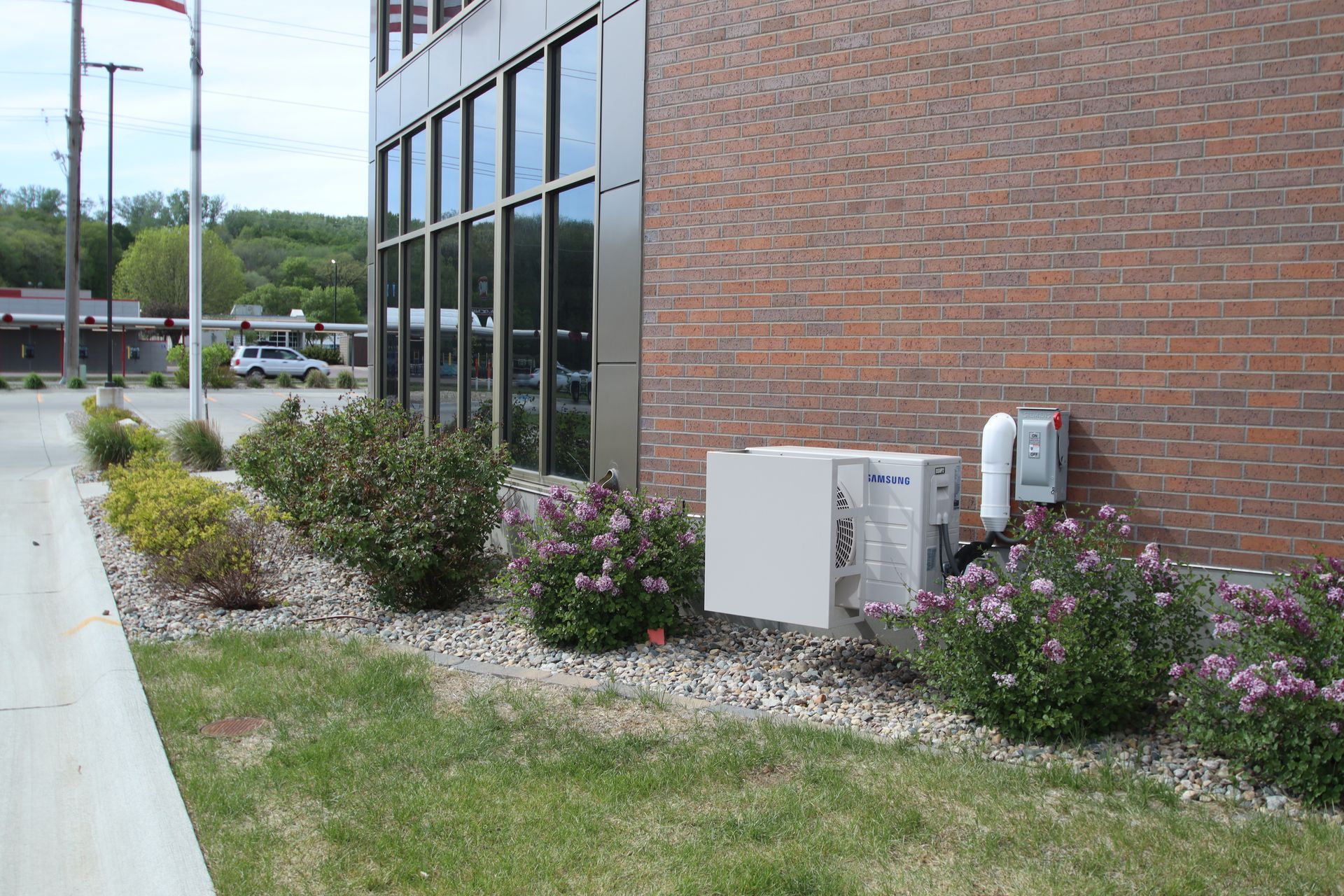 Brick building side wall with windows, utility boxes, and flower shrubs along a grassy edge.