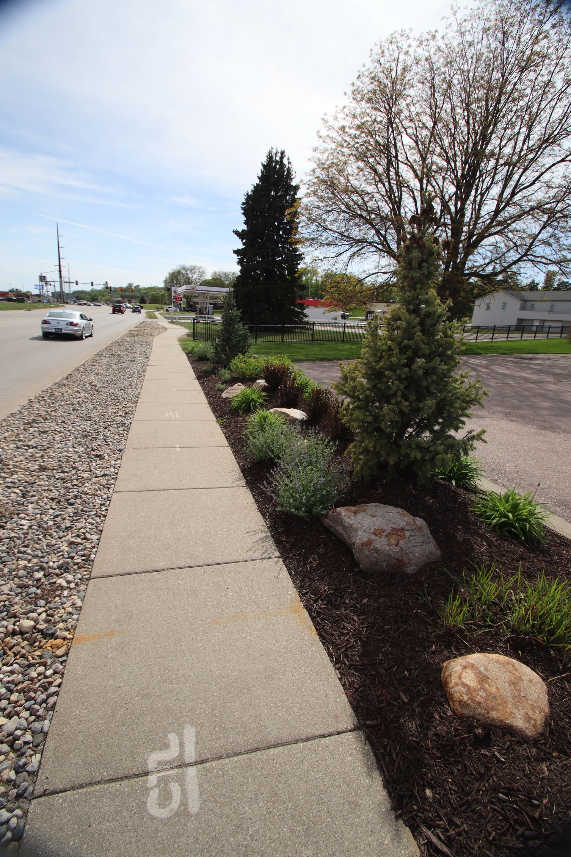 Sidewalk beside a gravel road with landscaped shrubs, rocks, and trees on an overcast day.