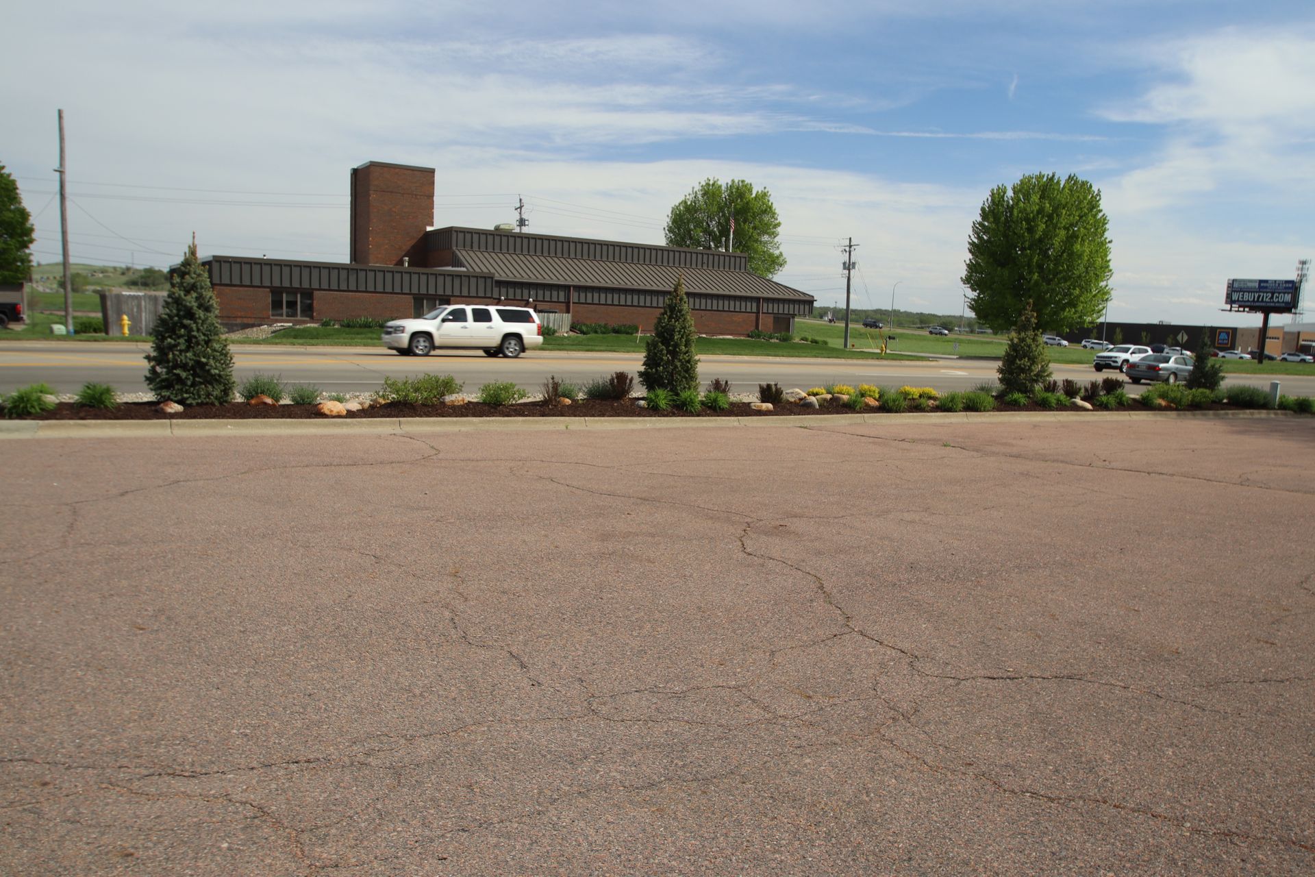 Parking lot with a white SUV, shrubs, and a low brick building under a clear sky.