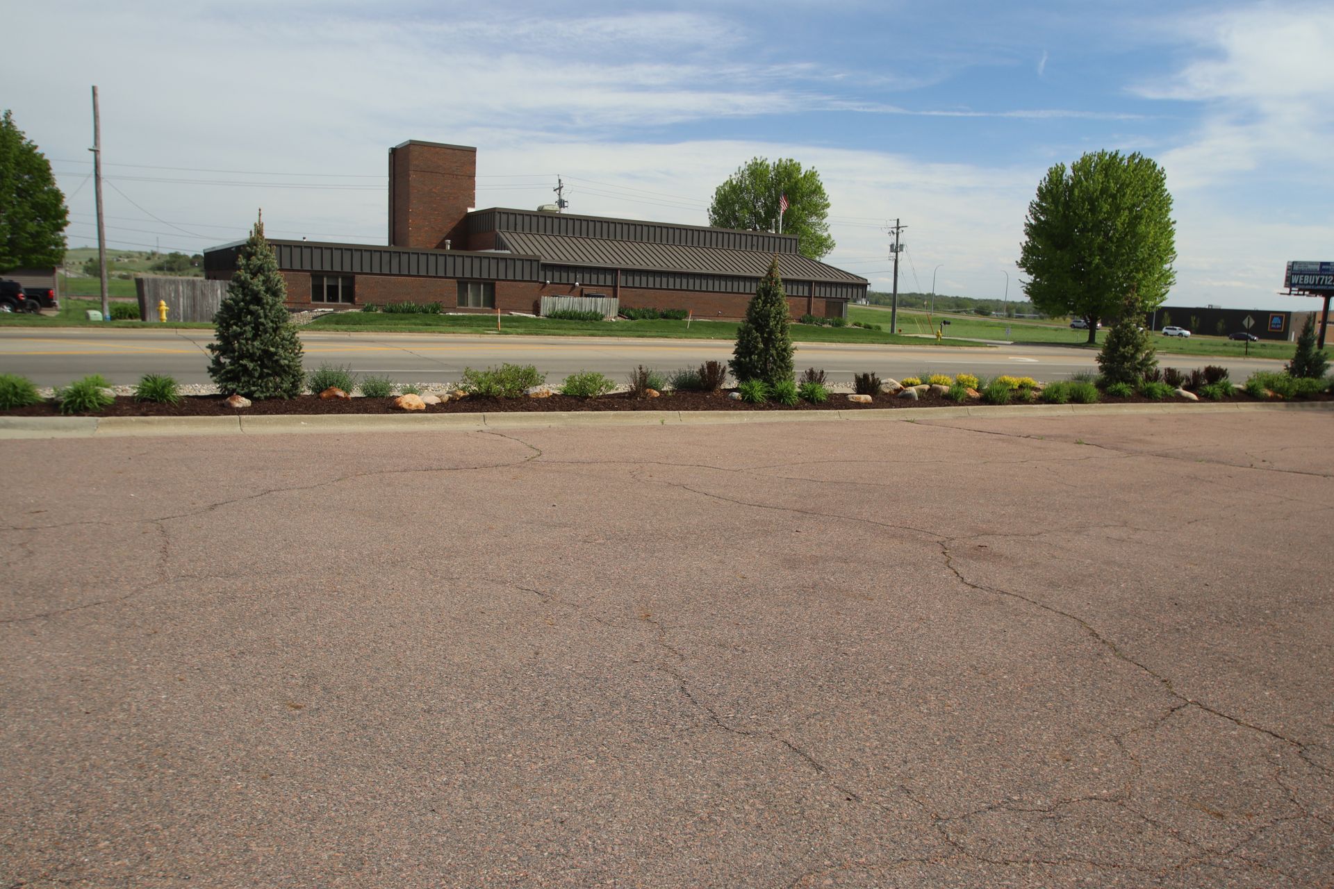 Empty parking lot with landscaped median and a low brick building under a partly cloudy sky