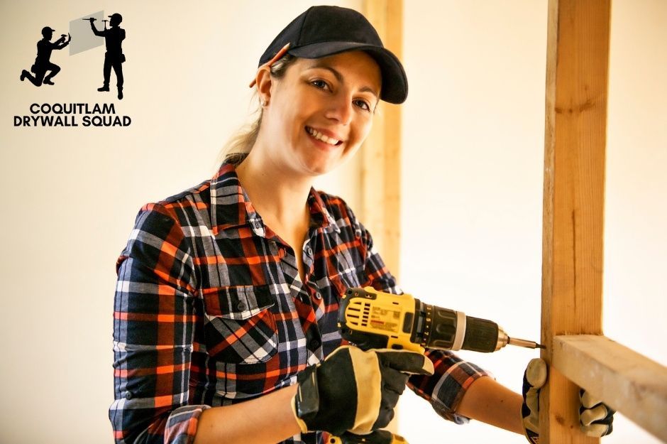 A skilled drywall repair company worker smiles while operating a yellow drill during a commercial renovation project. She wears a black cap and plaid flannel shirt, working among exposed wooden framing near Buena Vista Ave, and Kent St in White Rock, BC.