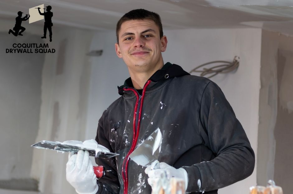 A professional drywall repair handyman wearing a dark hoodie and work gloves holds a putty knife with compound during a commercial renovation project. He stands confidently in an unfinished interior space with white walls near Gifford St, and Basran Ave in New Westminster, BC.