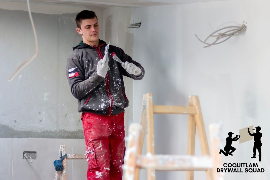 A professional drywall repair specialist wearing red work pants uses a trowel for the residential walls. He stands on the flooring with a wooden ladder nearby, applying paint finish during interior renovation work near Glencoe Dr, and Glenayre Dr in Port Moody, BC.