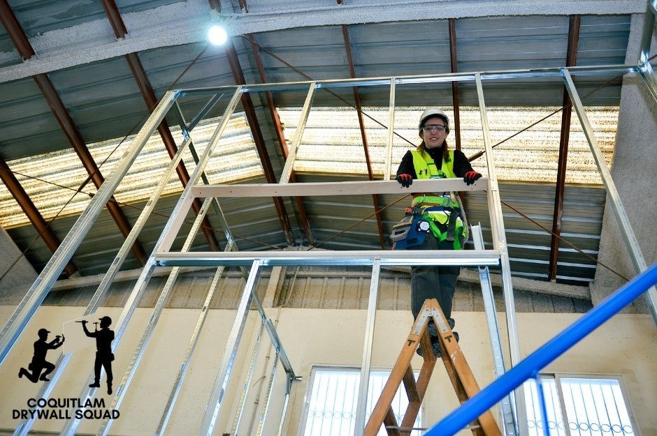A professional drywall repair specialist works on metal framing from an elevated platform in a commercial construction project. She wears a bright safety vest and a hard hat while installing or adjusting a ceiling framework at Southarm in Richmond, BC.