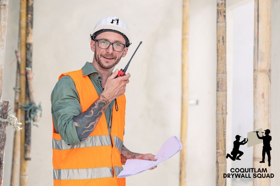 A professional drywall repair technician wearing glasses, a hard hat, and an orange vest holds papers and a walkie-talkie while inspecting a residential construction site. He smiles confidently among exposed wooden wall studs near Marine Dr, and Royal Oak Ave in Burnaby, BC.