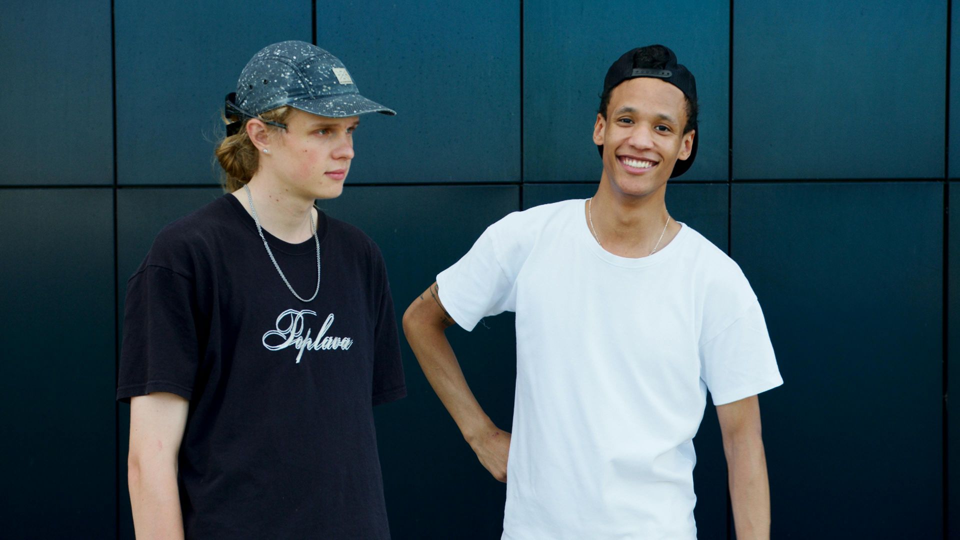 Two young people standing by a dark wall, one in a black shirt and cap, the other in a white T-shirt, smiling.