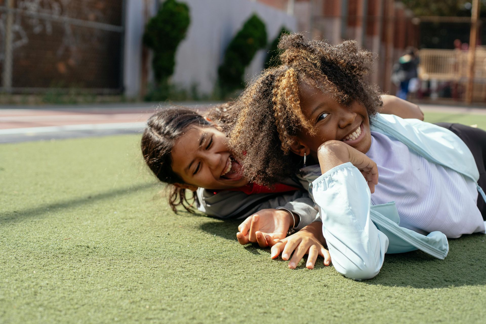 Two children lying on green turf, smiling and resting their heads on their hands outdoors.