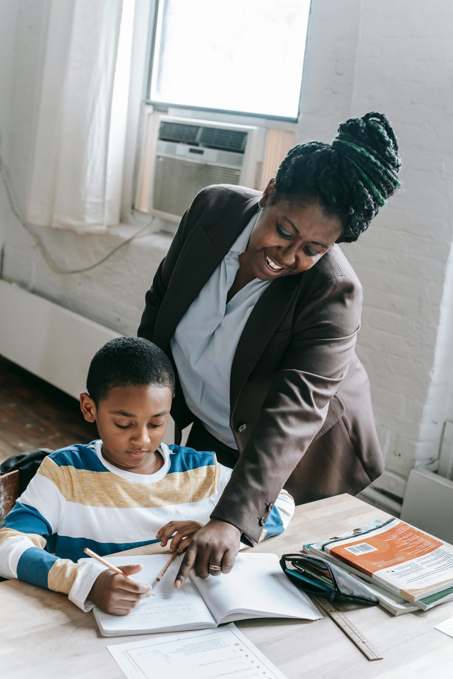 Teacher helping a child study at a desk with open books and papers near a window.