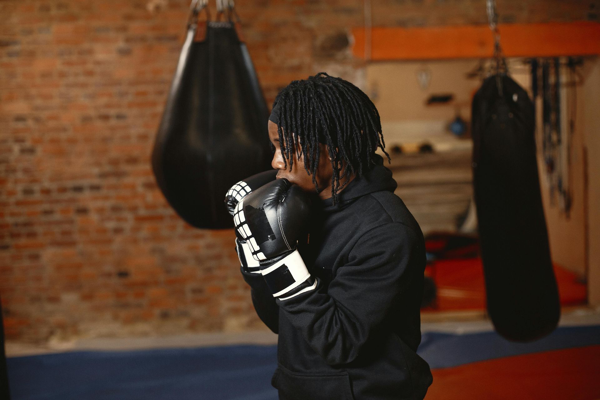 Person in boxing gloves training in a gym with hanging punching bags and a brick wall.