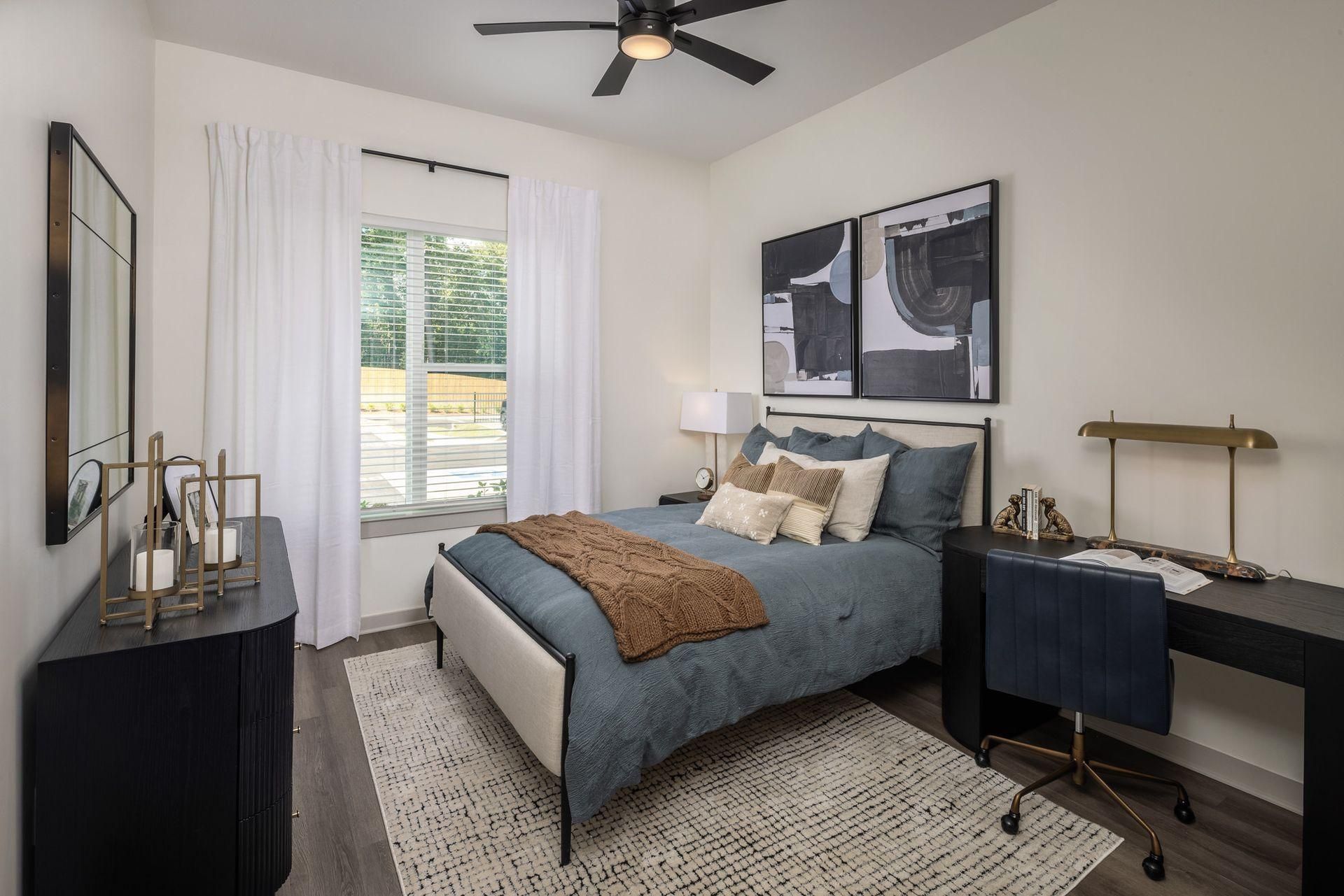 Cozy bedroom with a bed, desk, lamp, wall art, and a window with white curtains.