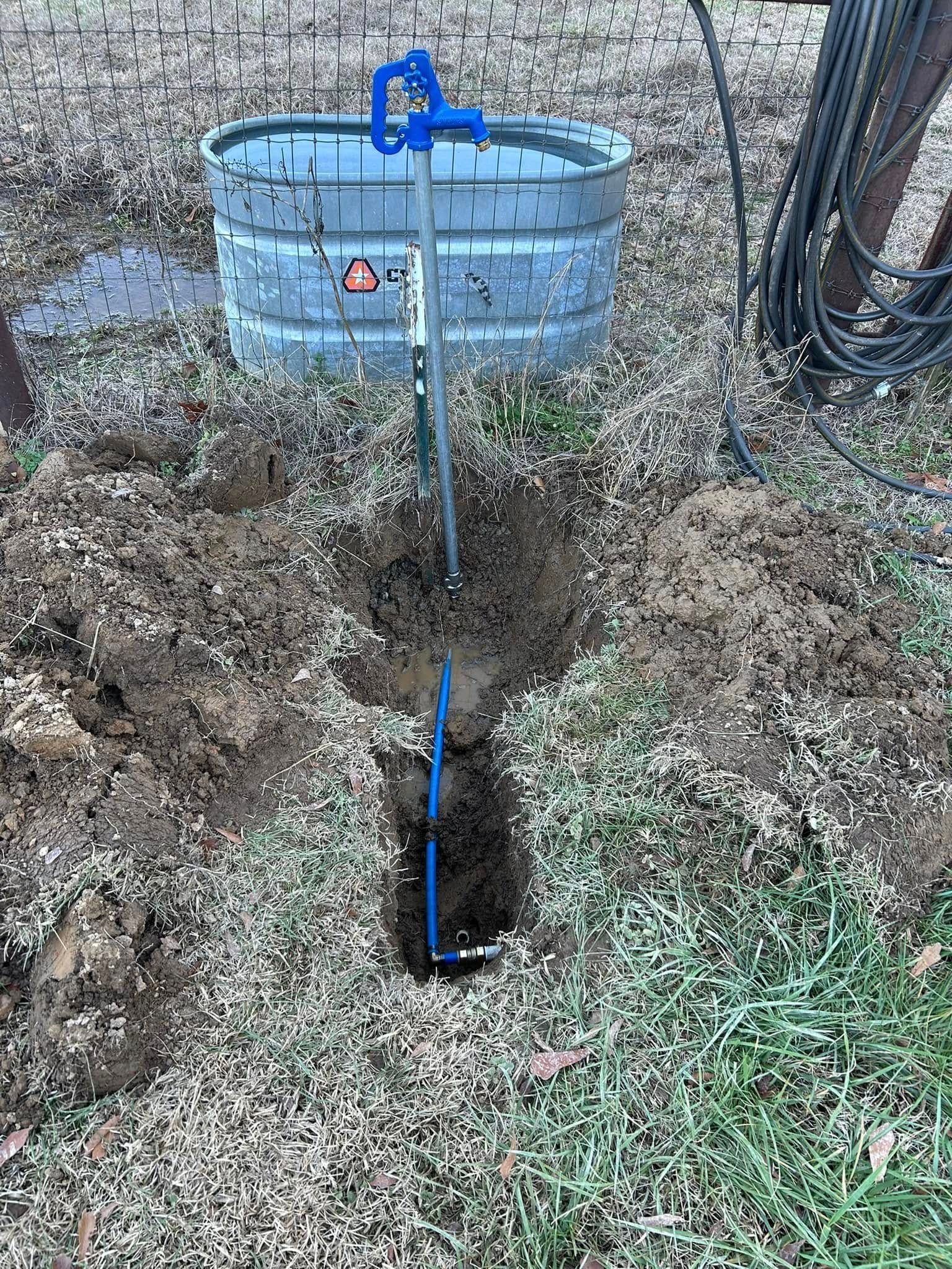 A blue water hydrant installed in a dirt trench, connected to a blue pipe, next to a galvanized metal water trough.