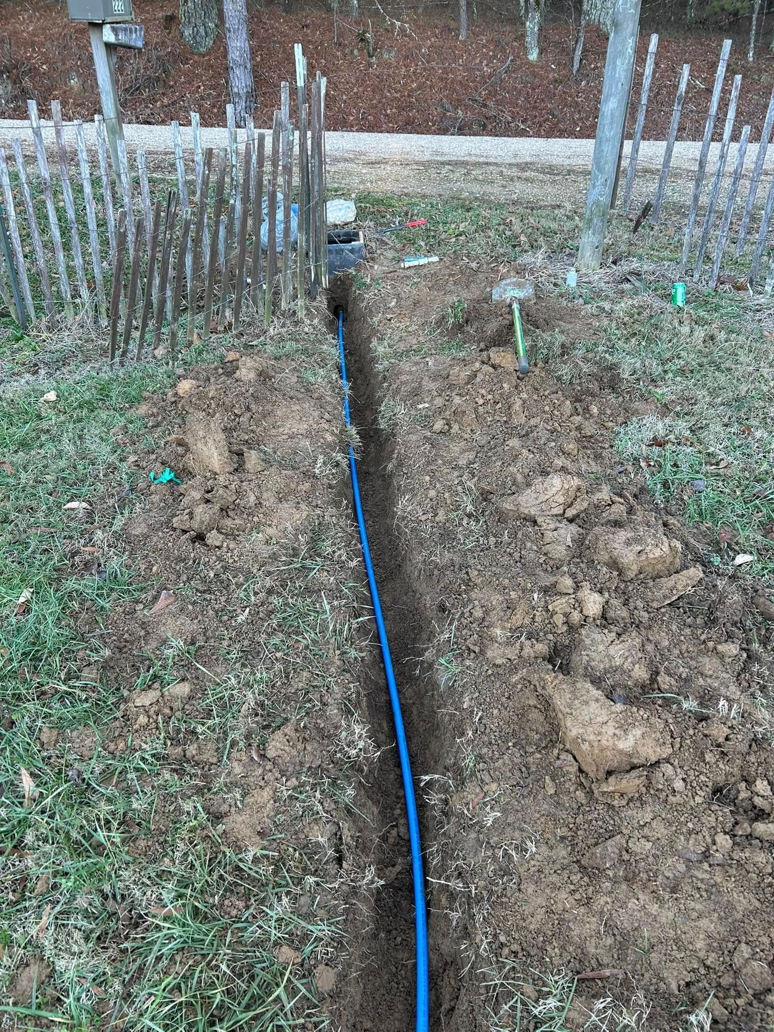 A blue irrigation pipe runs through a narrow, dug-out trench in a residential yard next to a wooden fence.