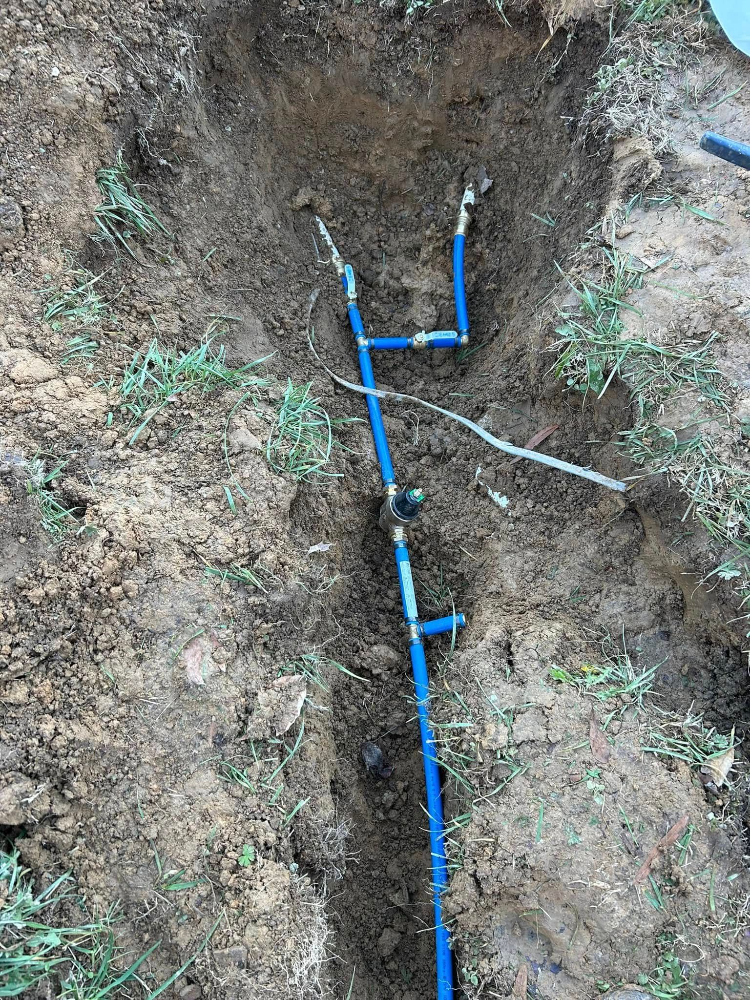 Blue PEX pipe segments connected with brass fittings and elbows installed in a narrow dirt trench in the ground.