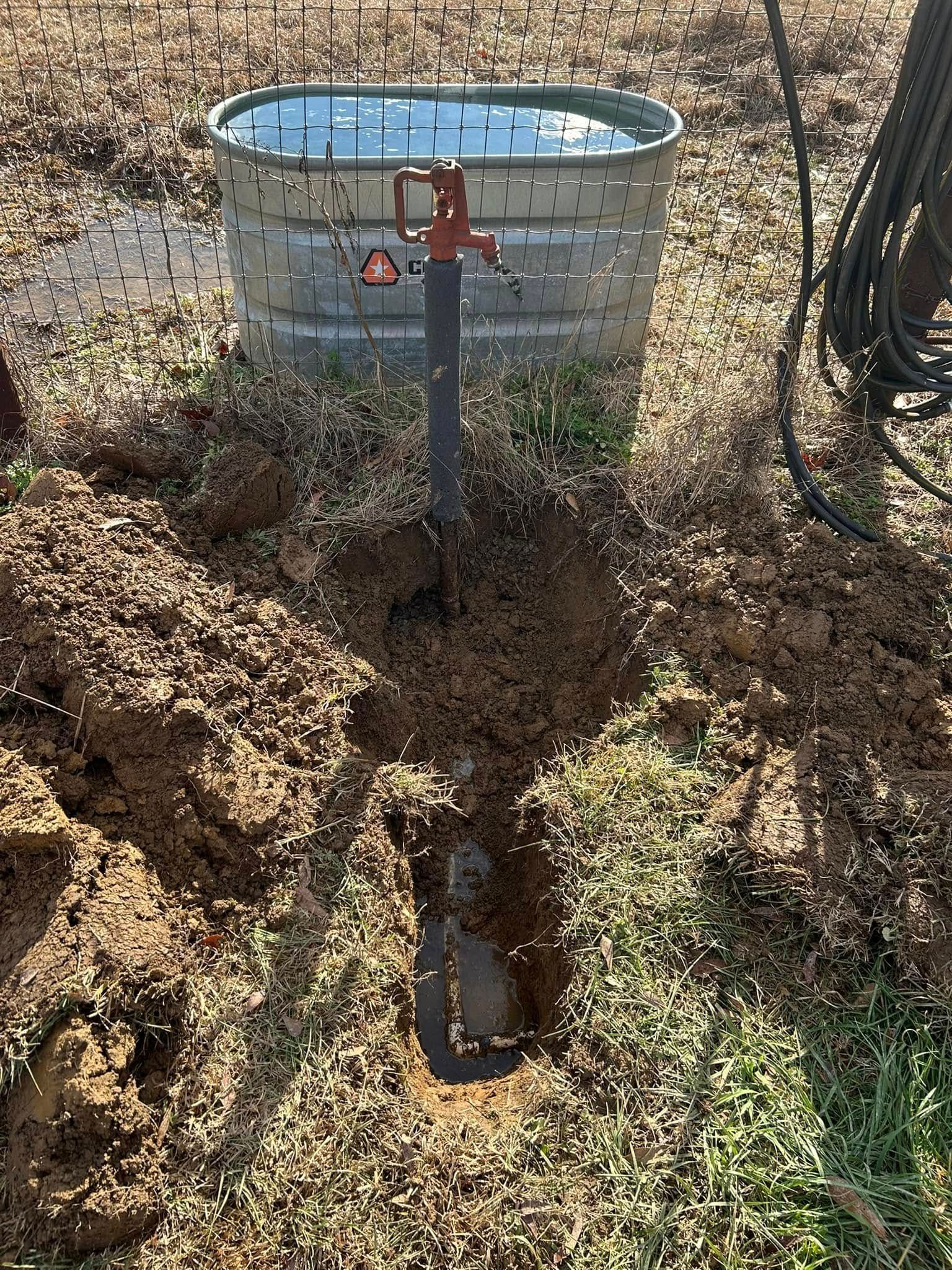 A metal livestock water trough stands in a dirt field next to a vertical water pump pipe installed in a shallow trench.