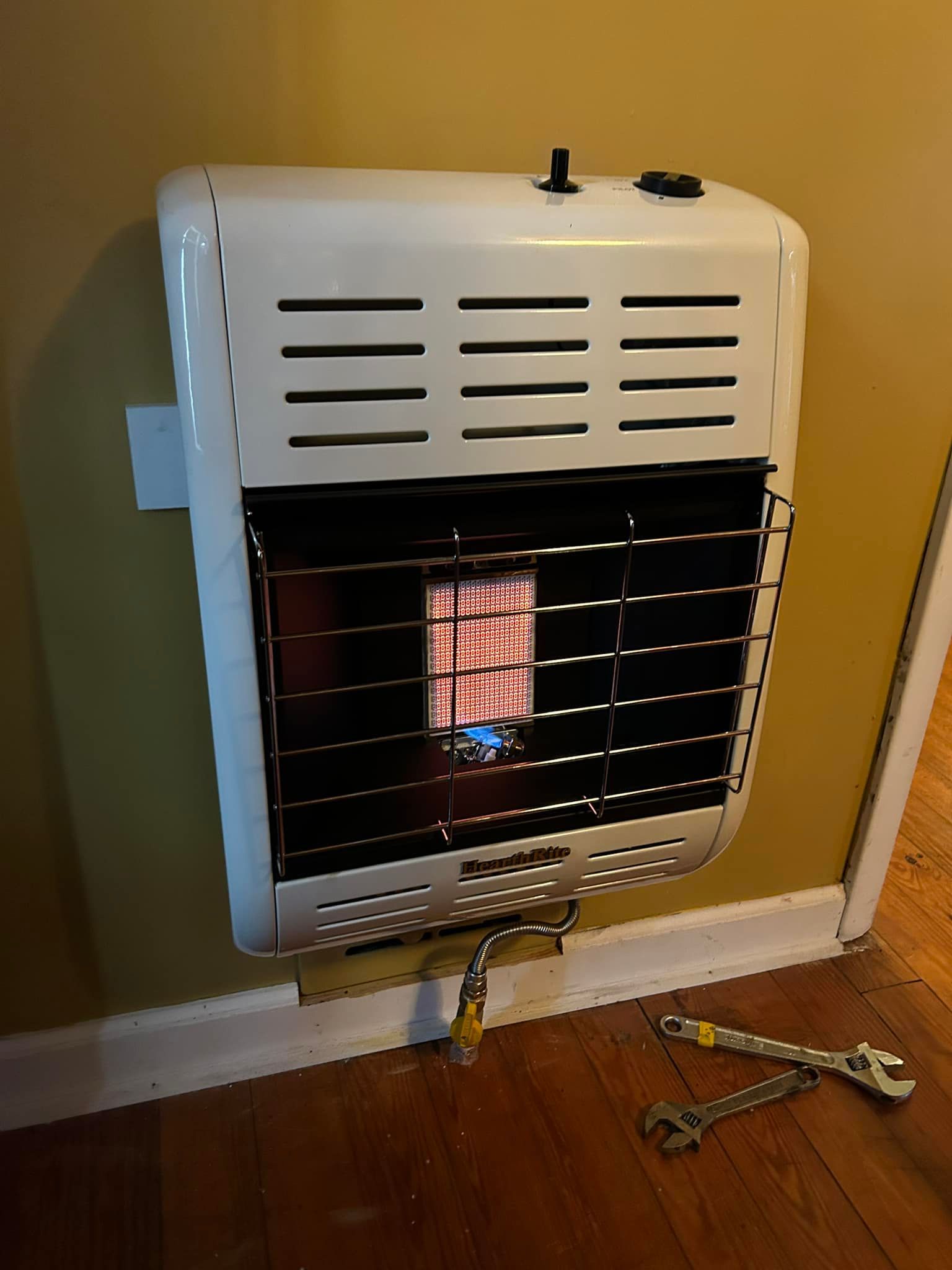 A white wall-mounted gas heater with a glowing ceramic heating element, located near two wrenches on a wooden floor.