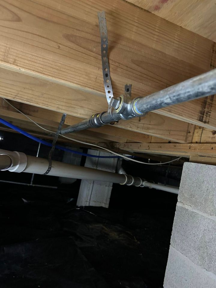 Metal gas pipe hung from a wooden floor joist by a perforated metal plumbing strap in a dark crawlspace.