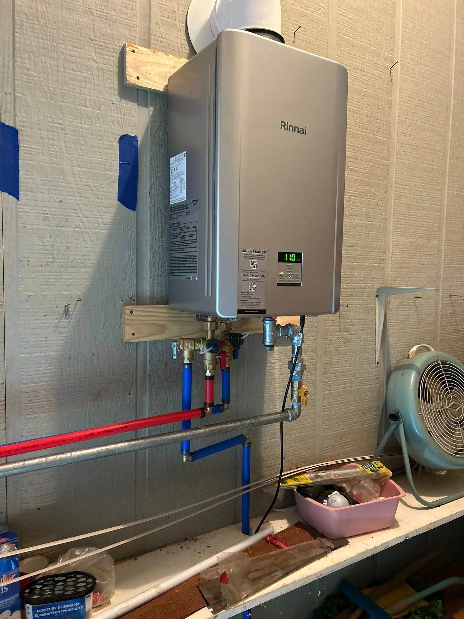 A silver tankless water heater mounted on a wooden frame against a light-colored wall with visible blue and red piping.