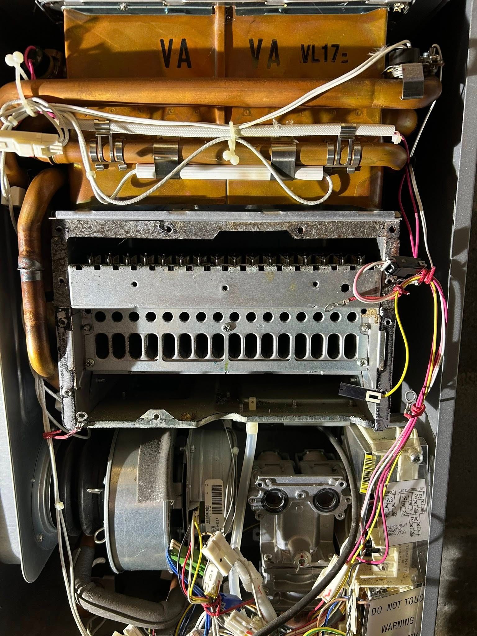 Internal view of a wall-mounted gas boiler with exposed copper pipes, heat exchanger, burner assembly, and electrical wires.