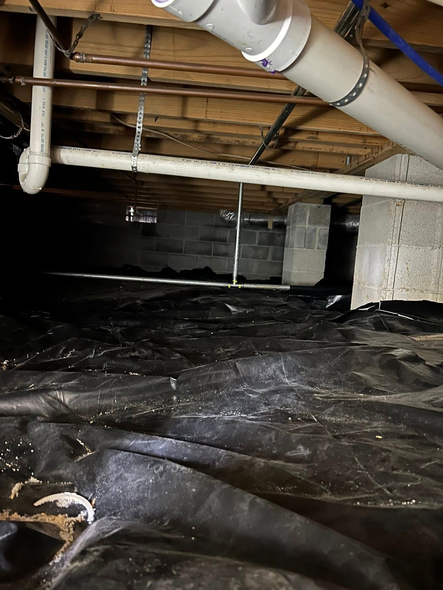 A view under a building floor showing PVC pipes, wooden joists, concrete block supports, and a black plastic ground cover.