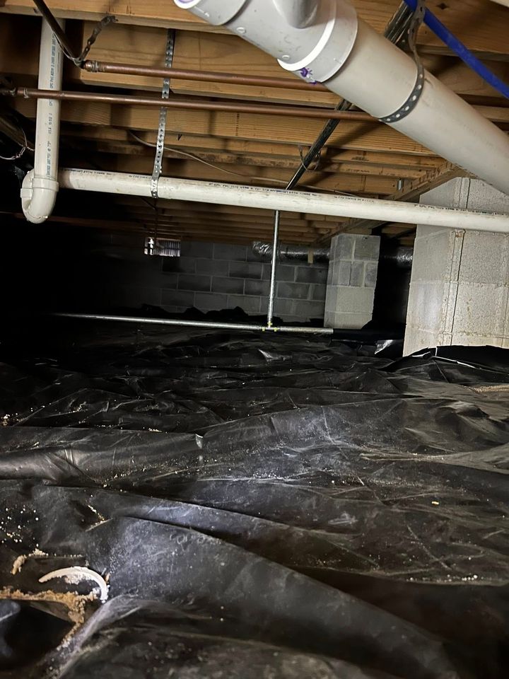 A view under a building floor showing PVC pipes, wooden joists, concrete block supports, and a black plastic ground cover.