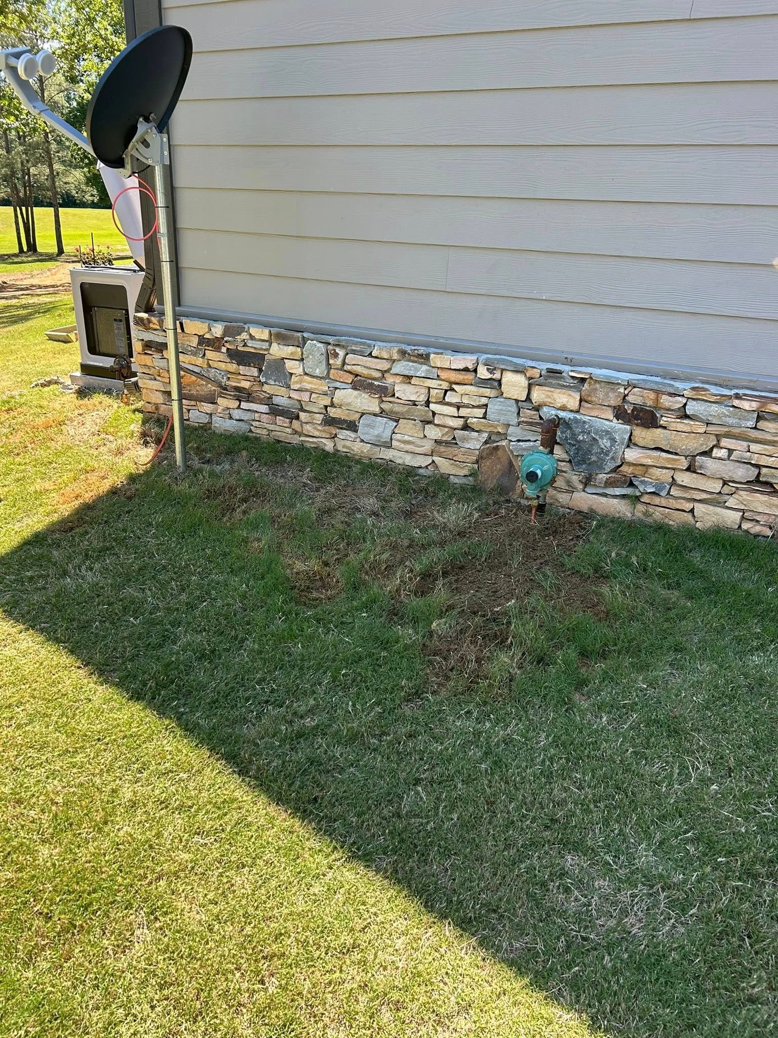 A satellite dish mounted to the side of a house with stone skirting, situated above a patch of dirt in a grassy lawn.