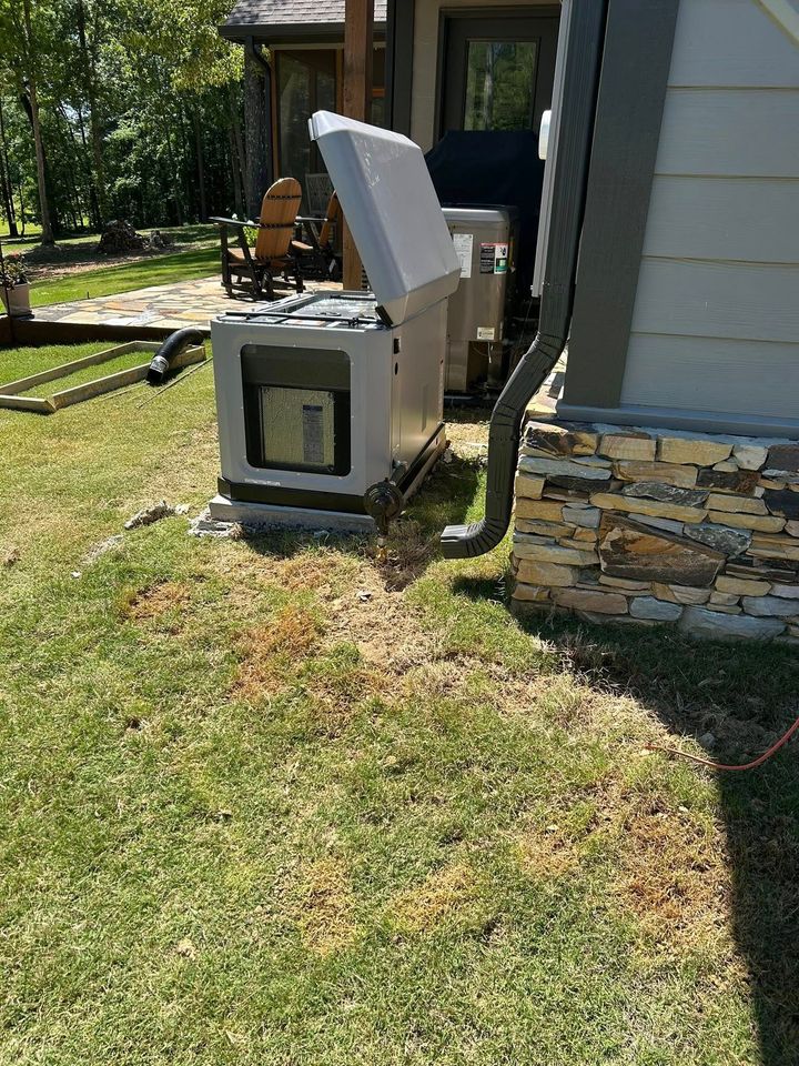 A light-colored residential standby generator with its lid open, installed in a grassy yard next to a stone house foundation.