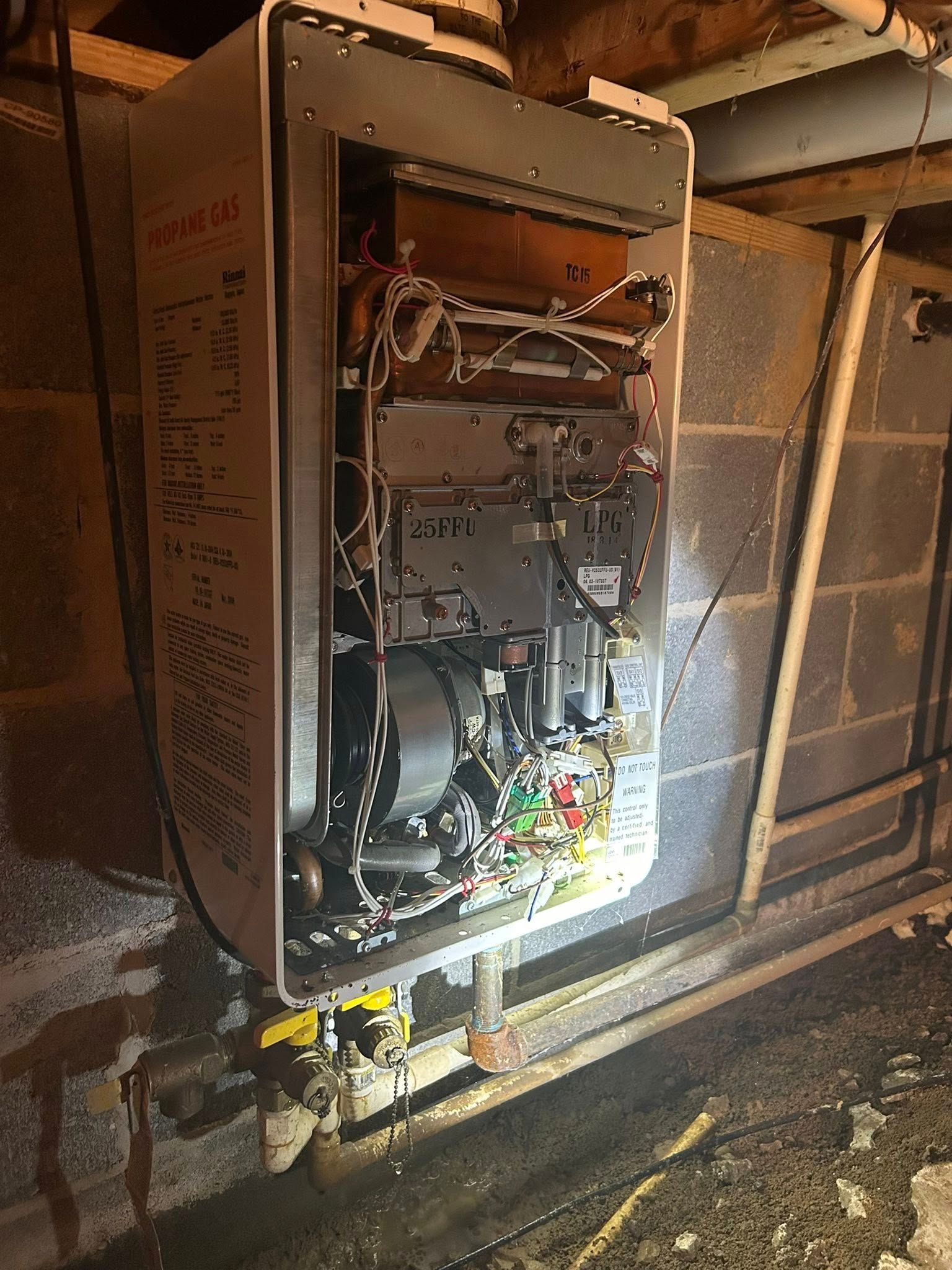 A wall-mounted, open-cased tankless water heater with visible internal wiring and components in a crawl space.