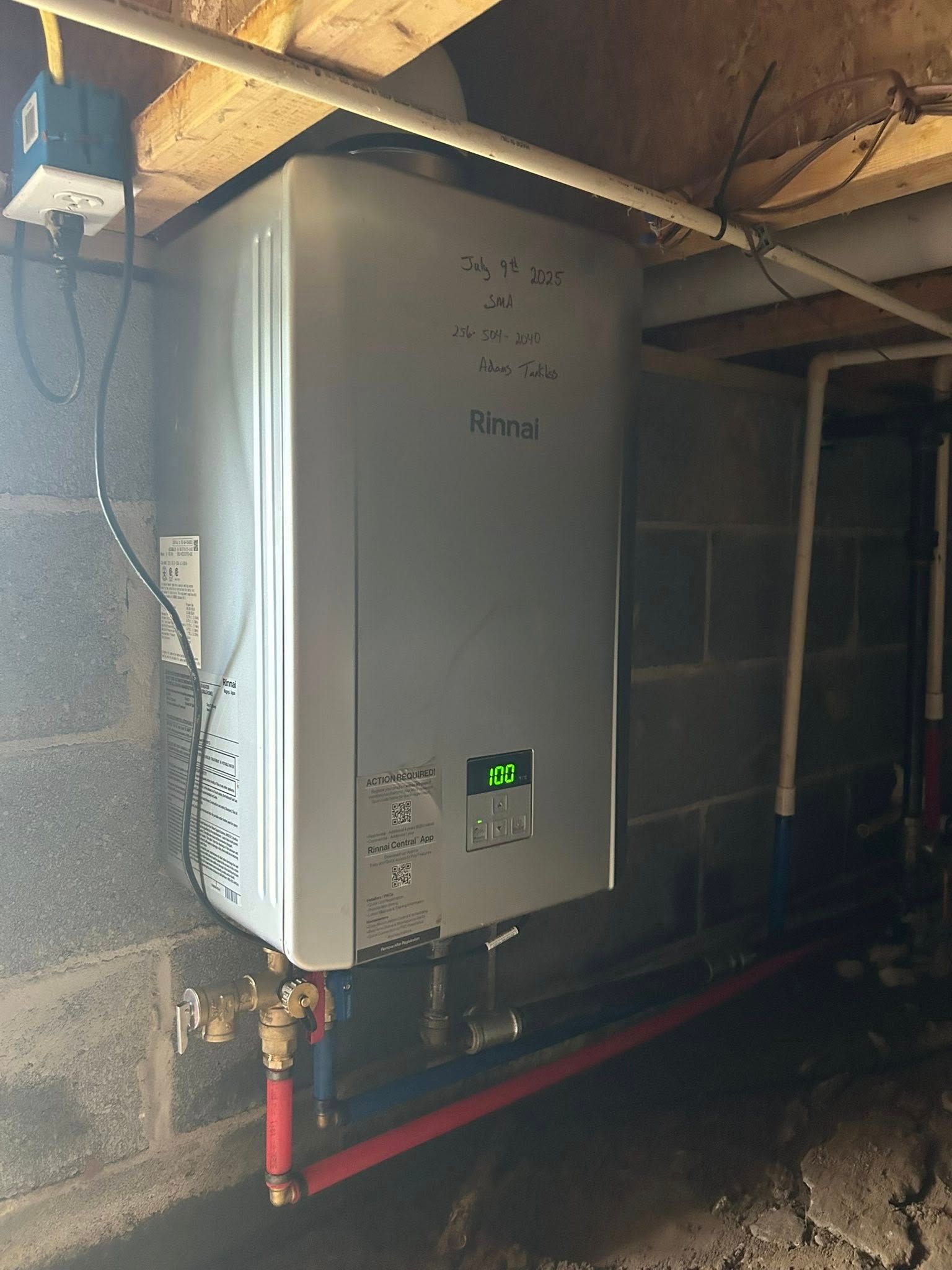 A silver tankless water heater mounted on a concrete block wall in a basement, connected to red and white pipes.