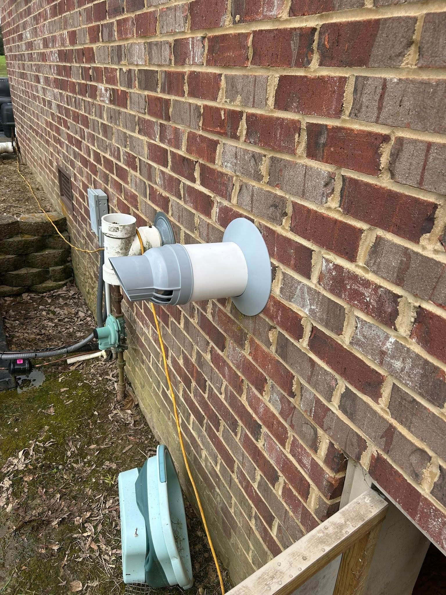 A white PVC exhaust vent pipe with a grey cover protruding from an exterior brick wall near a basement window.