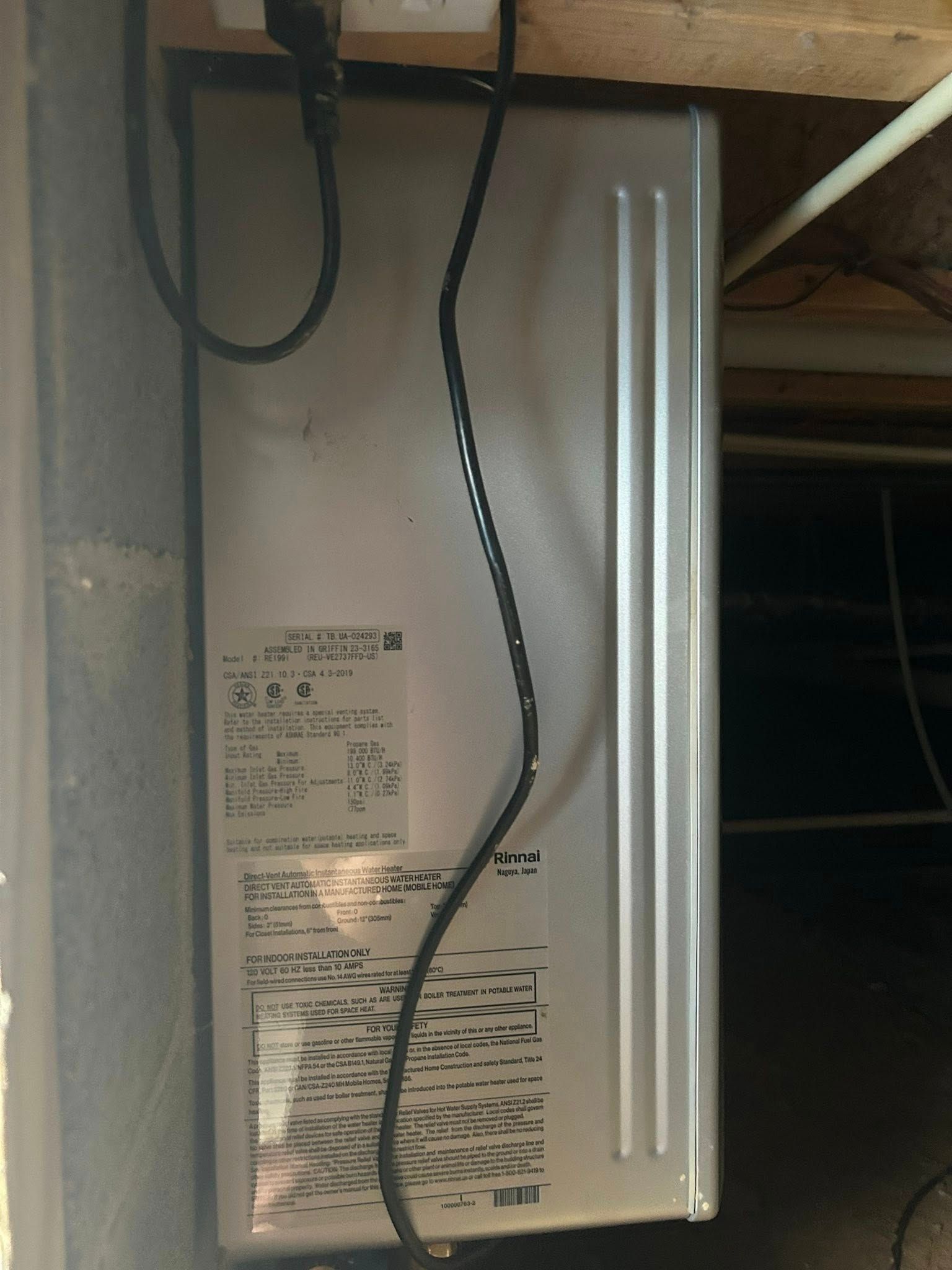 A gray metal electrical box or appliance mounted on a wall in a basement or crawlspace with a power cord plugged in above.