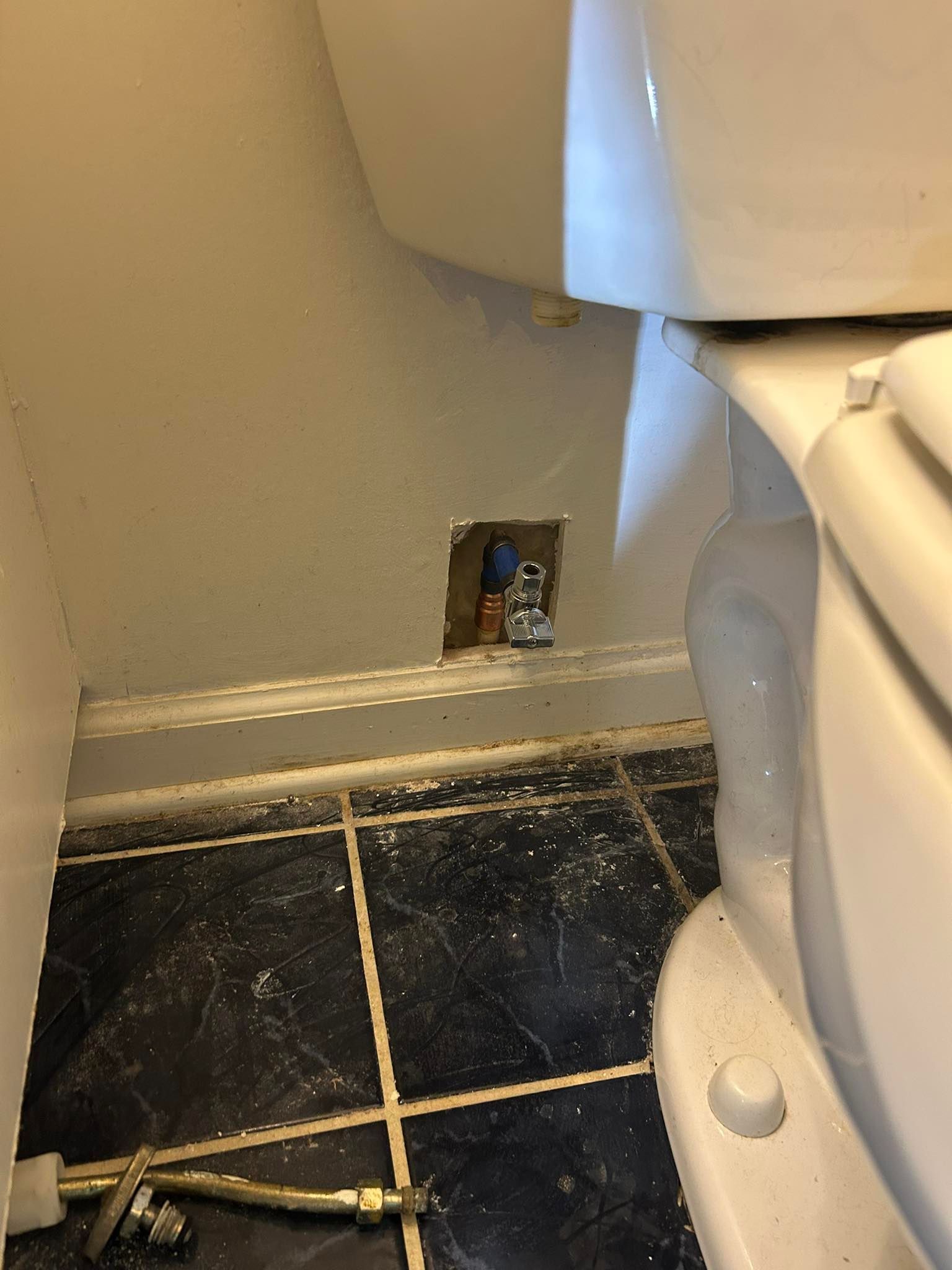 A detached toilet water supply hose lies on a dark tiled floor next to a bathroom wall with an exposed plumbing shutoff valve.