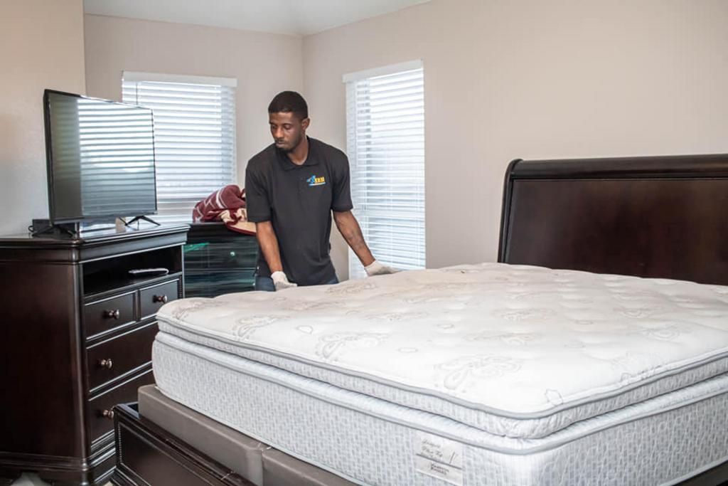 Person moving a mattress in a bedroom, with a dresser, TV, and blinds.