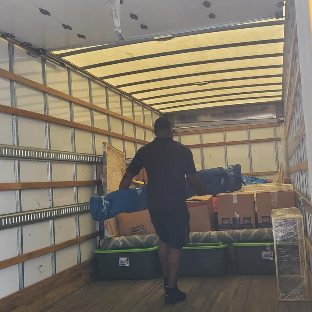 Person carrying a rolled-up blue object into a moving truck filled with boxes and containers.