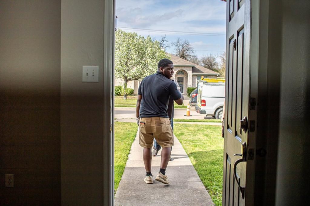 Man stepping outside doorway; white van parked on the lawn; trees and house in the background.