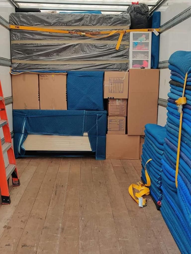 Inside a moving truck packed with furniture, boxes, and protective blankets. A ladder is on the left.