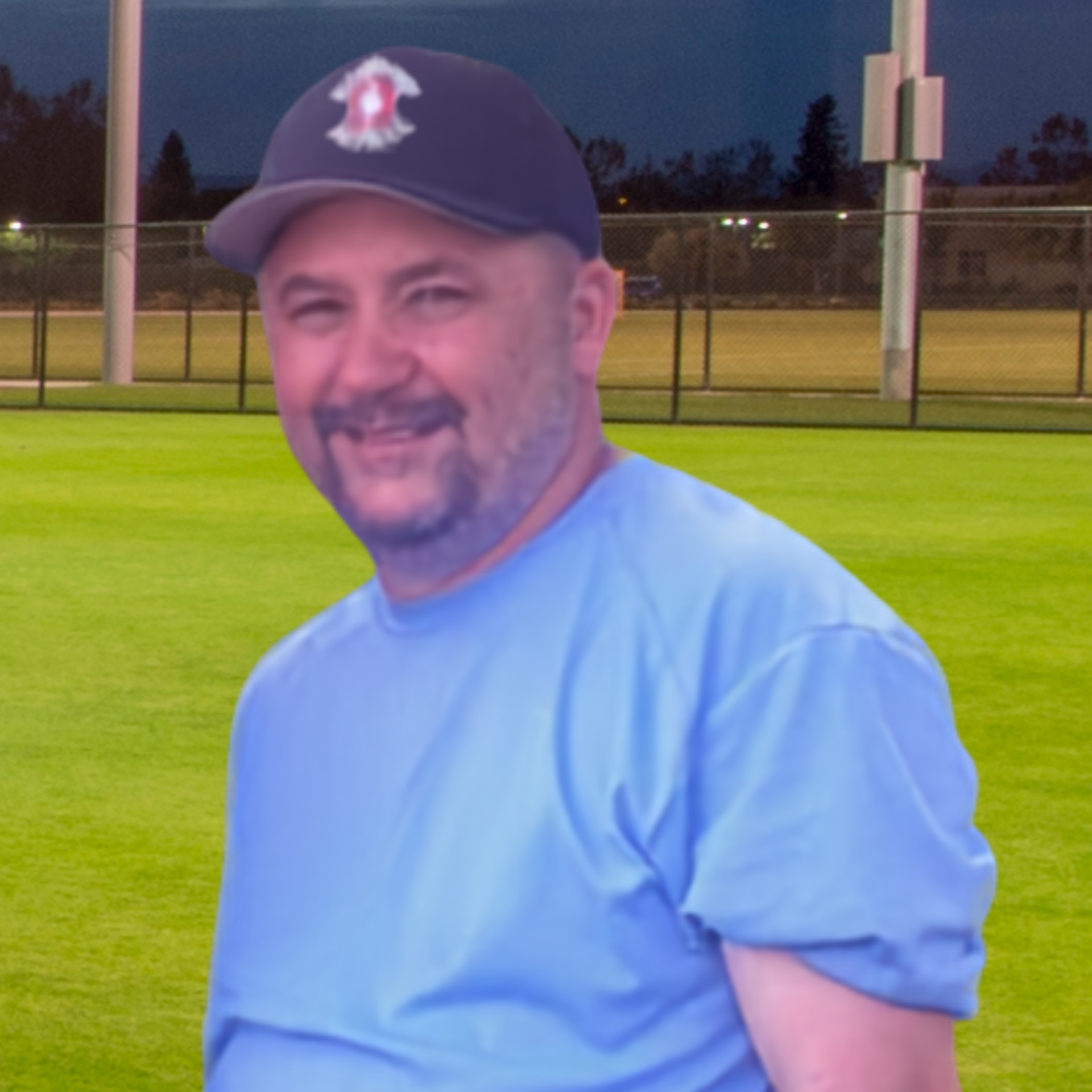 Smiling man in a blue T-shirt and black cap standing on a lit sports field at dusk
