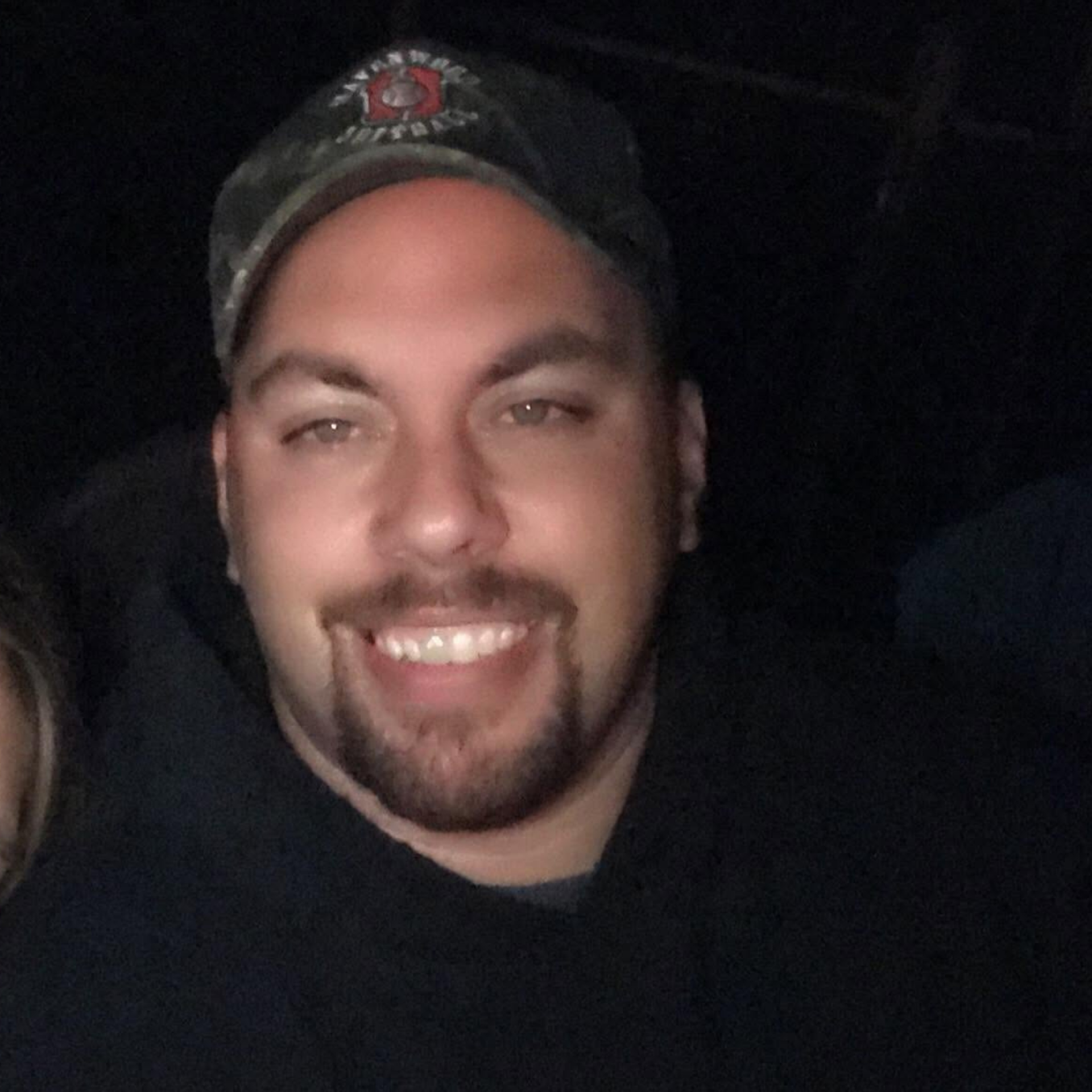 Smiling man in a dark cap and shirt, lit by flash against a dark background.