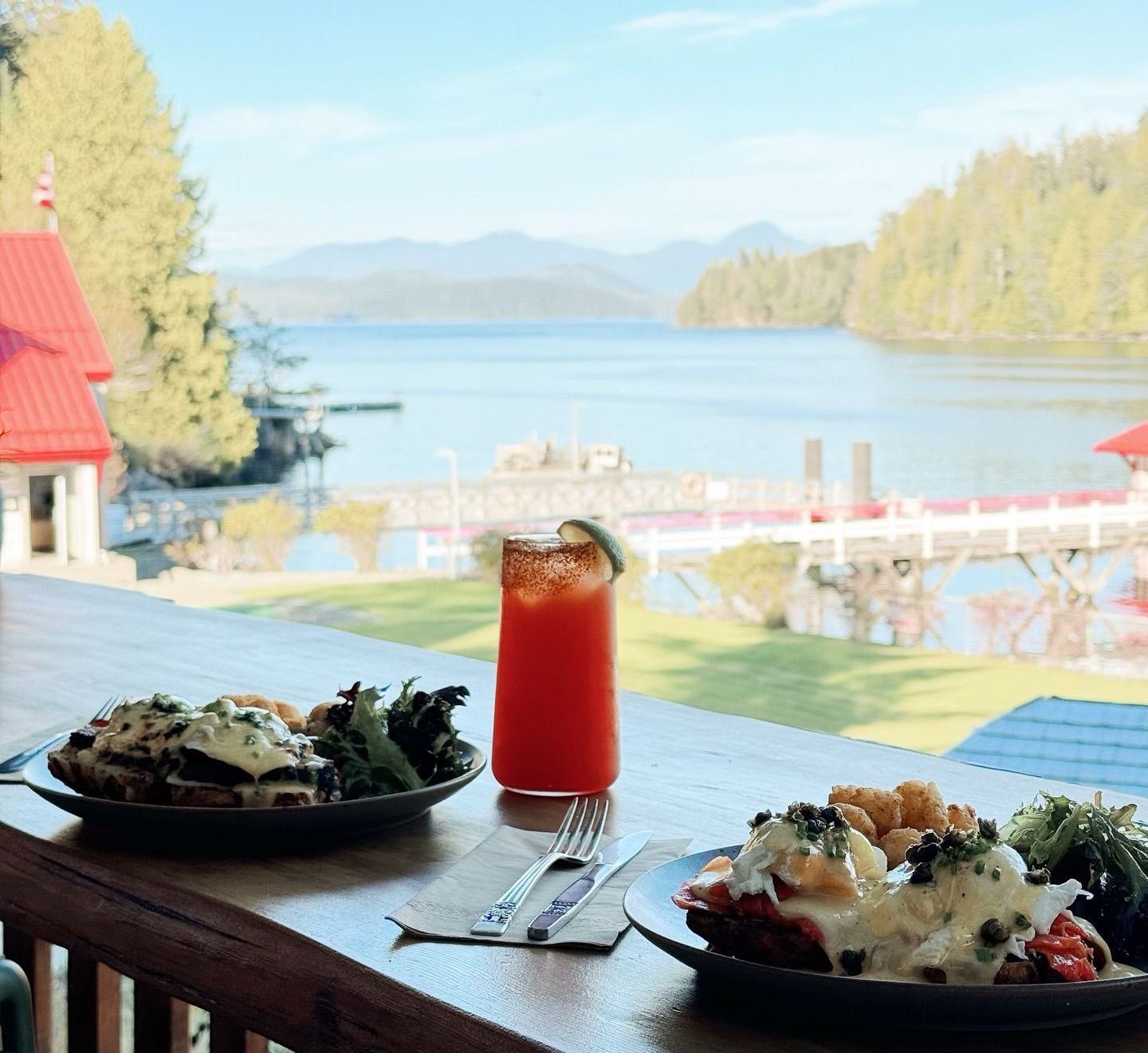 Two plates of eggs Benedict and a red drink on a wooden table, overlooking a scenic lake and mountains.