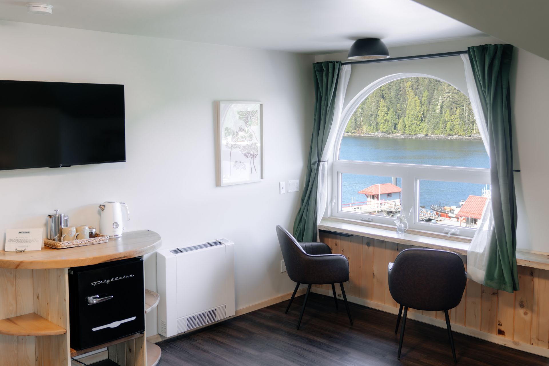 A hotel room features a television, a small fridge, two chairs, and a large arched window overlooking a water view.